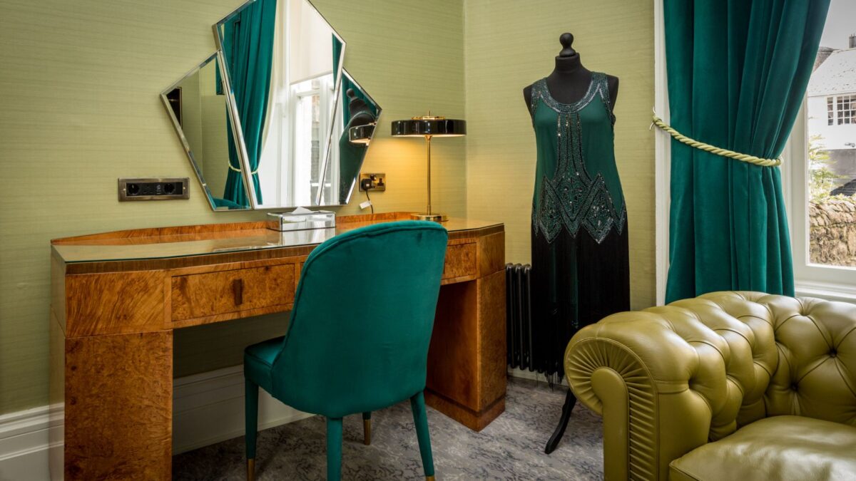 Luxurious hotel room with elegant vanity, teal chair, vintage dress display, and plush leather sofa by a sunlit window.