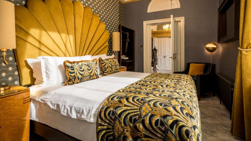 Luxurious hotel room with art deco décor, plush yellow headboard, patterned bedding, and warm lighting for cosy relaxation.
