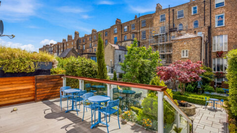 Charming hotel terrace with blue chairs, lush garden, and historic brick buildings, perfect for a serene outdoor retreat.