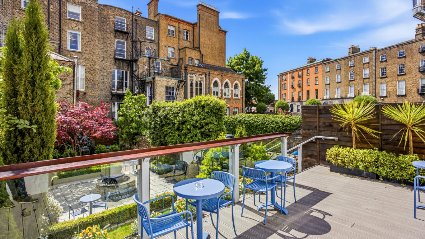 Charming garden view from terrace with blue chairs and tables, perfect for relaxing. Historic architecture surrounds lush greenery.